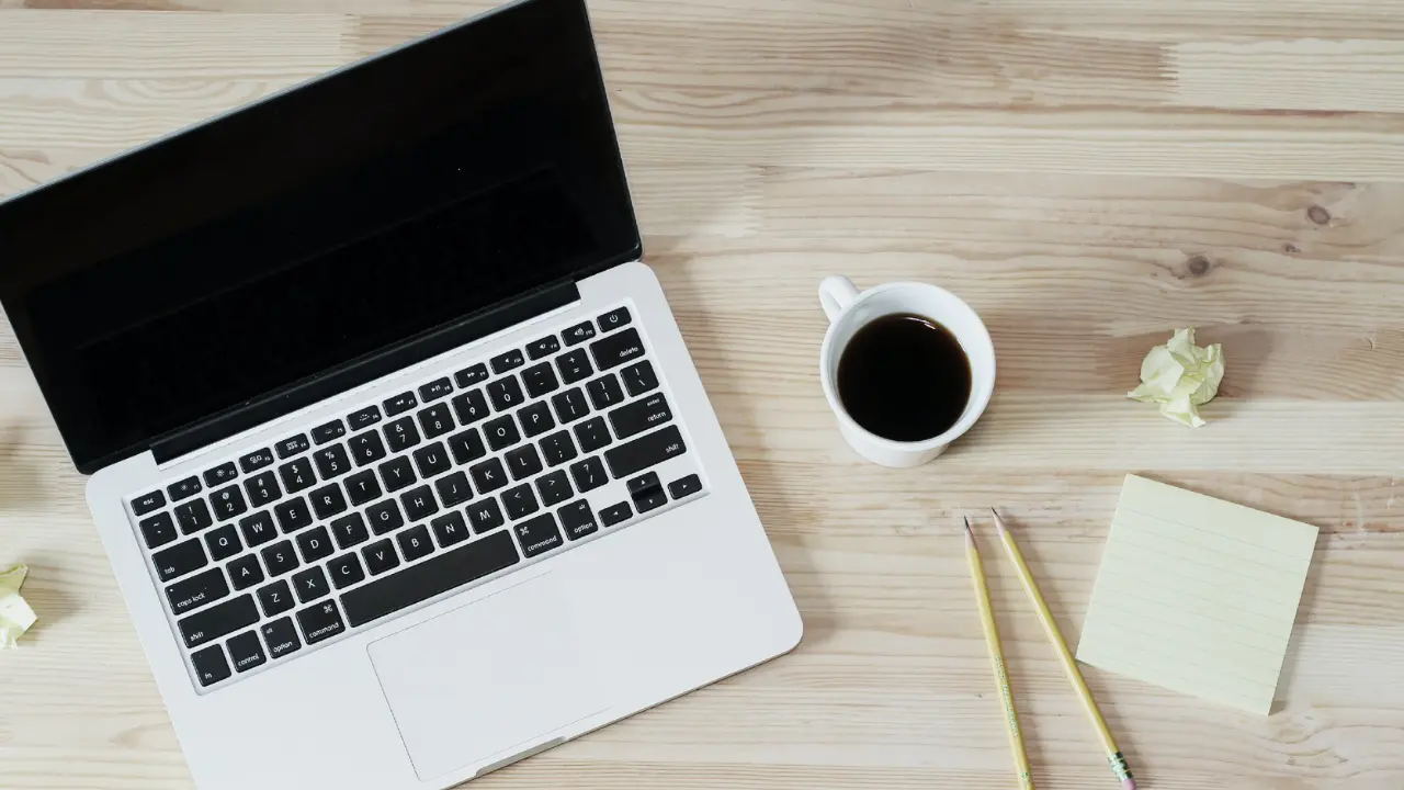 A laptop, sticky notes, pencils, crumpled notes, and a black coffee sitting on a light wooden surface.